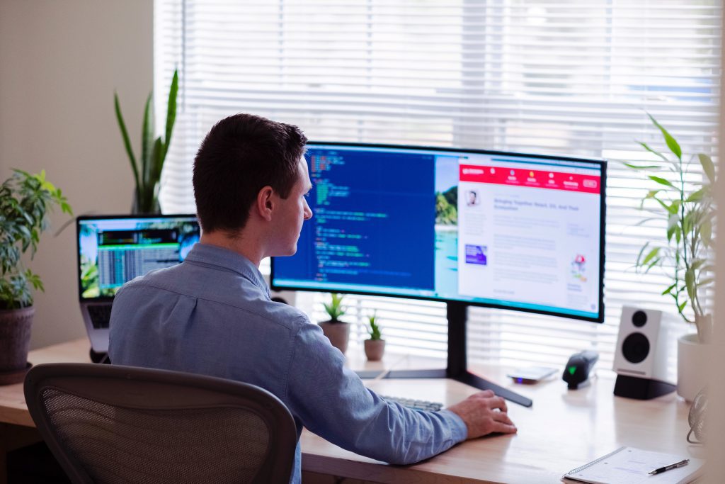 Man in a blue collared shirt sitting at a desk with plants with three computer monitors and evaluating lease accounting software platforms.